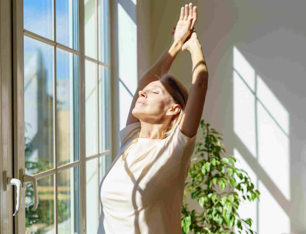 Well-rested mature woman stretches before window in the morning.